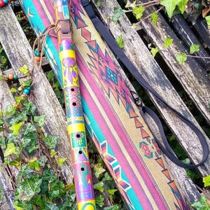 May include: A cylindrical wooden flute featuring a vibrant yellow, red, and green painted finish with geometric patterns, a lizard motif, and a brown mouthpiece, accompanied by a matching fabric carrying case with a colourful woven design.