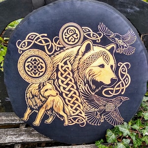 May include: A circular black cushion with a golden wolf design, Celtic knotwork, and two ravens. The wolf is the central image, with detailed knot patterns and a smaller wolf figure. The design is set against a dark background.
