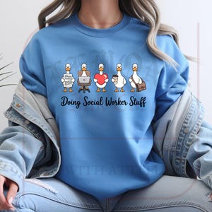May include: A light blue sweatshirt with the phrase "Doing Social Worker Stuff" in black script. The graphic features five cartoon ducks, each with a different item: paperwork, a laptop, a heart, a coffee cup, and a messenger bag.