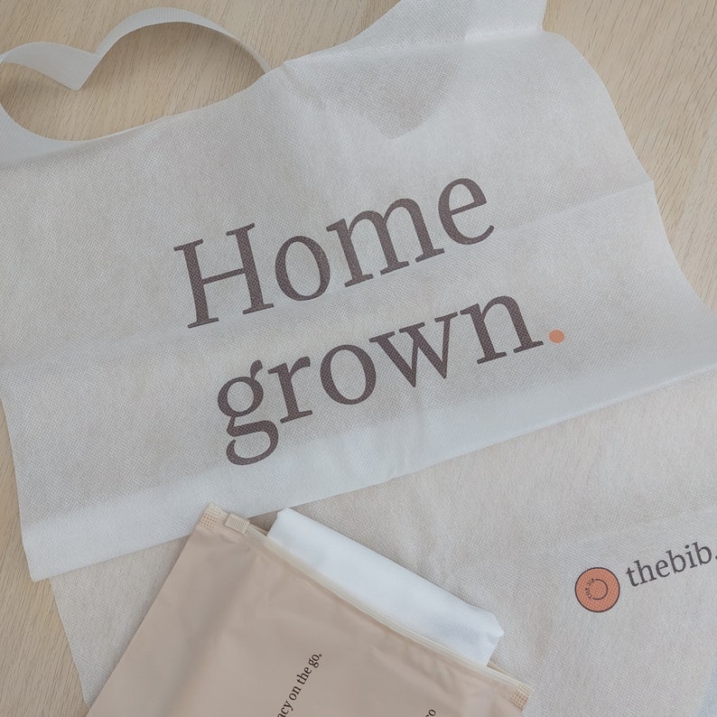 May include: White reusable cloth bib with the text "Home grown." printed in brown. The bib is folded in half and has a small brown pouch with the text "thebib" and a circle logo printed on it.