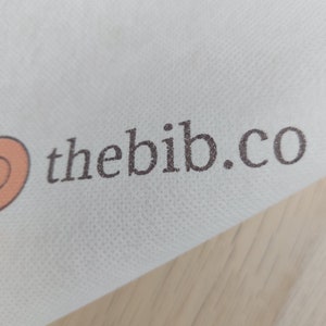 May include: White fabric with the text "thebib.co" printed in brown.