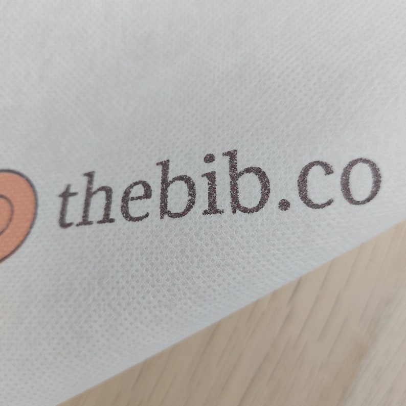 May include: White fabric with the text "thebib.co" printed in brown.