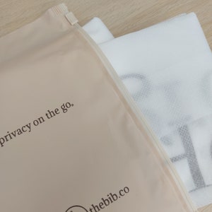 May include: A beige zippered pouch with the text "privacy on the go." and the website "thebib.co" printed on the front. The pouch is partially open, revealing a white mesh fabric.