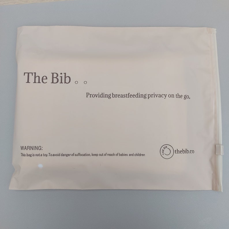 May include: A white mailing bag with the text "The Bib" and the tagline "Providing breastfeeding privacy on the go." The bag also has a warning label that reads "This bag is not a toy. To avoid danger of suffocation, keep out of reach of babies and children." The bag has a zipper and a logo that reads "thebib.co".