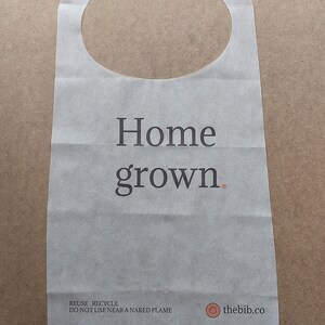 May include: White paper bib with the text "Home grown." printed in black. The bib has a round neck opening and the text "REUSE. RECYCLE. DO NOT USE NEAR A NAKED FLAME" printed in black at the bottom. A small circle with the text "thebib.co" is printed in brown on the right side of the bib.