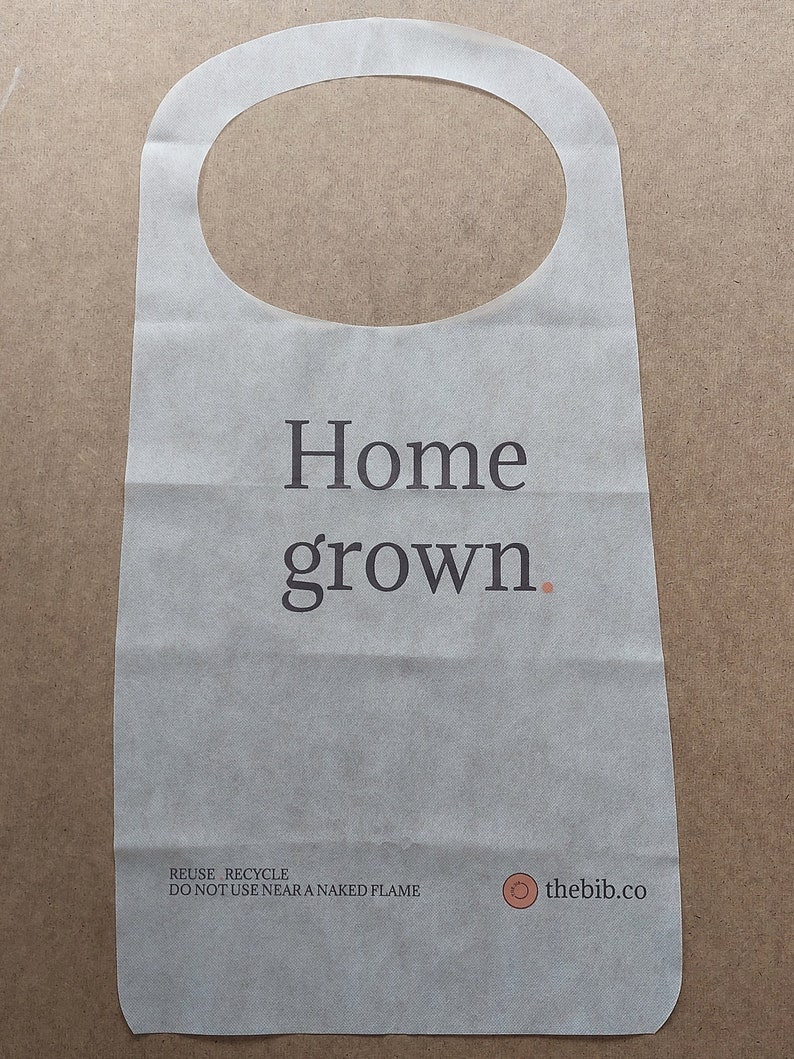 May include: White paper bib with the text "Home grown." printed in black. The bib has a round neck opening and the text "REUSE. RECYCLE. DO NOT USE NEAR A NAKED FLAME" printed in black at the bottom. A small circle with the text "thebib.co" is printed in brown on the right side of the bib.
