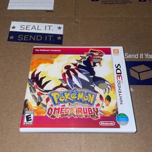 Puede incluir: Caja de juego de Nintendo 3DS para "Pokémon Omega Ruby" con una criatura roja, blanca y negra en la portada. La caja del juego es blanca con el logotipo de Nintendo 3DS y la clasificación ESRB.
