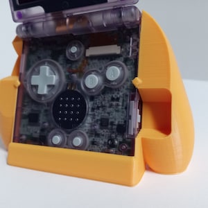 May include: An orange 3D-printed Game Boy Advance SP grip. The handheld console is visible inside the grip, with its transparent buttons and screen. The grip has a curved design for comfortable handling during gameplay.