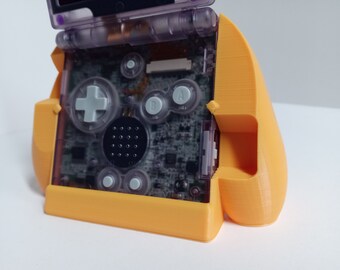 May include: An orange 3D-printed Game Boy Advance SP grip. The handheld console is visible inside the grip, with its transparent buttons and screen. The grip has a curved design for comfortable handling during gameplay.