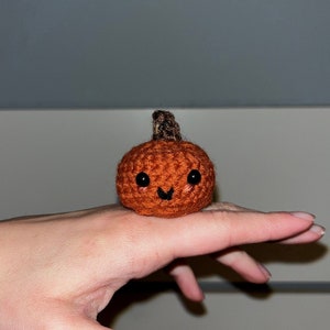 May include: A small crocheted pumpkin with a brown stem and a smiling face. The pumpkin is orange and has black eyes and a black mouth.