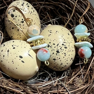 Op de afbeelding: Een paar oorbellen met blauwe en witte keramische kralen en gouden accenten. De oorbellen worden gepresenteerd in een vogelnest.