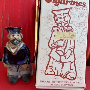 May include: A ceramic figurine of a bandsman wearing a dark blue uniform and a matching cap, accompanied by its original cardboard box. The box features the text "Figurines", "BANDSMAN", and "TREMAR POTTERIES (FIGURINES) LTD LISKEARD CORNWALL".