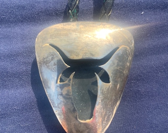 Op de afbeelding: Een zilveren bolo stropdas met een driehoekige hanger met een uitsparing van een stierenkop. De hanger hangt aan een zwart gevlochten leren koord, tegen een blauwe achtergrond.