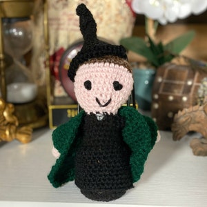 May include: A crocheted doll wearing a green robe and a black pointed hat. The doll has a pink face with black eyes and a smiling mouth.