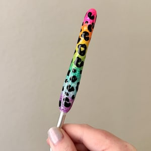 May include: A pen with a rainbow gradient design, transitioning from pink to purple, green, and yellow, adorned with black leopard spots. The pen has a silver metal base.