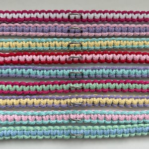 May include: Close-up of several colorful macrame dog collars. Each collar is a different color combination, including pink, purple, yellow, red, and blue. Each collar has a silver metal ring.