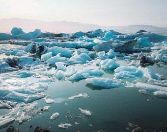 May include: A vast expanse of icy blue glacial icebergs floating in a turquoise lagoon. The icebergs are various shapes and sizes, with some appearing to be melting. The background is a hazy blue sky.