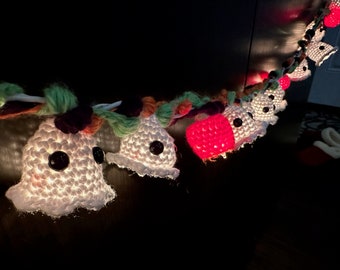 Può includere: Una ghirlanda di luci a crochet a forma di fantasmi e zucche. I fantasmi sono bianchi con occhi neri e le zucche sono rosse. Le luci sono infilate insieme con filato colorato.