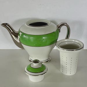 May include: A white ceramic teapot featuring a wide horizontal lime green band and metallic silver trim on the spout, handle, and base. The set includes a matching lid with a green band and a separate white cylindrical tea infuser with small holes.