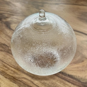 May include: A clear, spherical glass paperweight featuring a textured, frosted surface finish and a small, smooth, rounded knob at the top center. The object is transparent, allowing light to pass through its rounded form and intricate, uneven exterior.