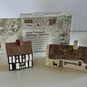 May include: Two ceramic miniature houses with detailed architectural features. One is black and white with a brown roof and a red chimney. The other is off-white with a thatched roof. The box reads "John Putnam's Heritage Houses."