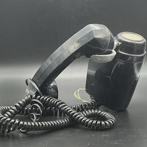 May include: A vintage black telephone handset with a coiled cord. The handset has a textured, angular design and a circular earpiece. The coiled cord is black and connects to the base of the phone. The phone is set against a dark background.