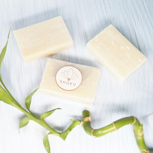 May include: Three rectangular bars of light beige soap are arranged on a pale blue surface. One bar features a circular sticker with the word "SHUKU" and a lotus flower design. Green bamboo stalks and leaves are in the foreground.