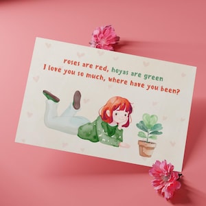 May include: A watercolor illustration on a card features a girl with red hair and a green sweater, lying down next to a potted plant. The text reads, "Roses are red, hoyas are green, I love you so much, where have you been?" Pink flowers are placed around the card.
