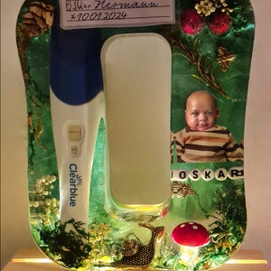 Può includere: Una cornice in resina verde con un test di gravidanza, una foto di un bambino e il testo "Oskar Herman *10.01.2024" su un'etichetta di carta bianca. La cornice è su una base di legno.