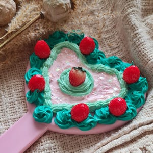 Könnte beinhalten: Ein rosa Handspiegel in Herzform. Er hat eine rosa Mitte mit grünem, frostingartigem Rand und roten Erdbeerdekorationen. Der Griff ist ebenfalls rosa.