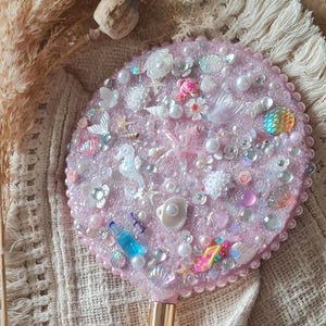 Könnte beinhalten: Ein runder, rosa Handspiegel, verziert mit maritimen Dekorationen. Der Spiegel hat Perlen, Muscheln, Seesterne und Miniatur-Seepferdchen. Der Griff ist hellrosa.