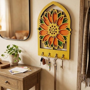 Op de afbeelding: Een gele houten sleutelhouder met een zonnebloemontwerp. De zonnebloem heeft oranje en groene details. De sleutelhouder heeft vier haken met sleutels en een hondenriem. Hij is bevestigd aan een crèmekleurige muur.
