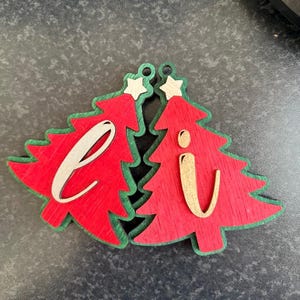 Op de afbeelding: Twee rode houten kerstboomhangers met groene randen, elk voorzien van een klein sterretje aan de bovenkant. De ene hanger toont de kleine letter l in licht hout, terwijl de andere de kleine letter i in licht hout toont.