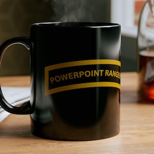 Könnte beinhalten: Eine schwarze Keramik-Tasse mit dem Text "POWERPOINT RANGER" in gelben Buchstaben. Die Tasse ist mit einem heißen Getränk gefüllt, und Dampf steigt von oben auf. Die Tasse steht auf einer Holzoberfläche.