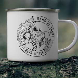 Peut inclure: Mug en émail blanc avec un dessin noir et blanc d'un crâne portant un casque, avec le texte "Hang in there" et "It gets worse". La tasse a un bord argenté et une anse blanche.