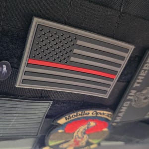 Può includere: Una collezione di toppe, tra cui una bandiera americana grigia e nera con una striscia rossa, un teschio e una toppa con le parole "Mobile Operations". Le toppe sono attaccate a una superficie nera.