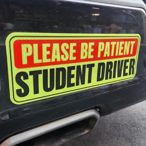 May include: A yellow and red student driver sign with the text "PLEASE BE PATIENT STUDENT DRIVER".