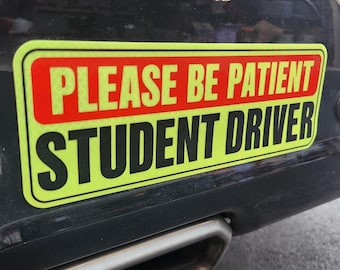 May include: A yellow and red student driver sign with the text "PLEASE BE PATIENT STUDENT DRIVER".