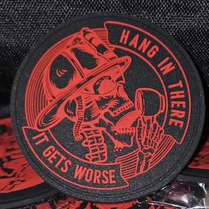 Peut inclure: Sous-verre rond noir et rouge avec un crâne portant un casque de pompier. Le texte sur le sous-verre est "Hang in there, it gets worse".