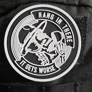 Pode incluir: Um patch redondo em preto e branco com as palavras "HANG IN THERE" e "IT GETS WORSE" circundando uma caveira com um chapéu. O patch é provavelmente feito de borracha ou material semelhante e está preso a uma superfície de tecido preto.