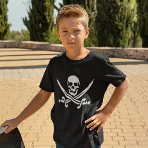 May include: A young person wearing a black t-shirt with a white skull and crossbones design, blue denim shorts, and white sneakers. They are holding a black skateboard with white lettering that says "The Skateboard".