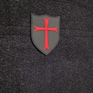 May include: A gray shield-shaped patch with a red cross emblem. The patch is on a dark gray textured fabric. Above the shield is a smaller patch with the text "WE'RE GOIN" in red.