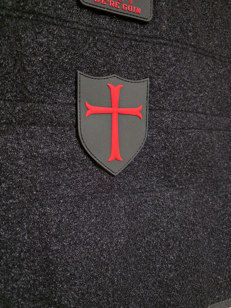 May include: A gray shield-shaped patch with a red cross emblem. The patch is on a dark gray textured fabric. Above the shield is a smaller patch with the text "WE'RE GOIN" in red.
