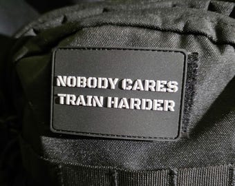 Può includere: Una toppa rettangolare nera con la scritta bianca "NOBODY CARES TRAIN HARDER". La toppa ha un bordo nero ed è attaccata a un tessuto nero strutturato. Probabilmente per una borsa o uno zaino.