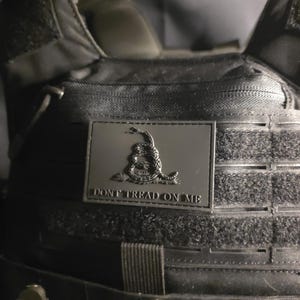 May include: Black tactical vest with a rectangular patch showing a coiled snake and the words "DON'T TREAD ON ME". The vest has multiple straps and pockets, and is made of a durable, heavy-duty material.