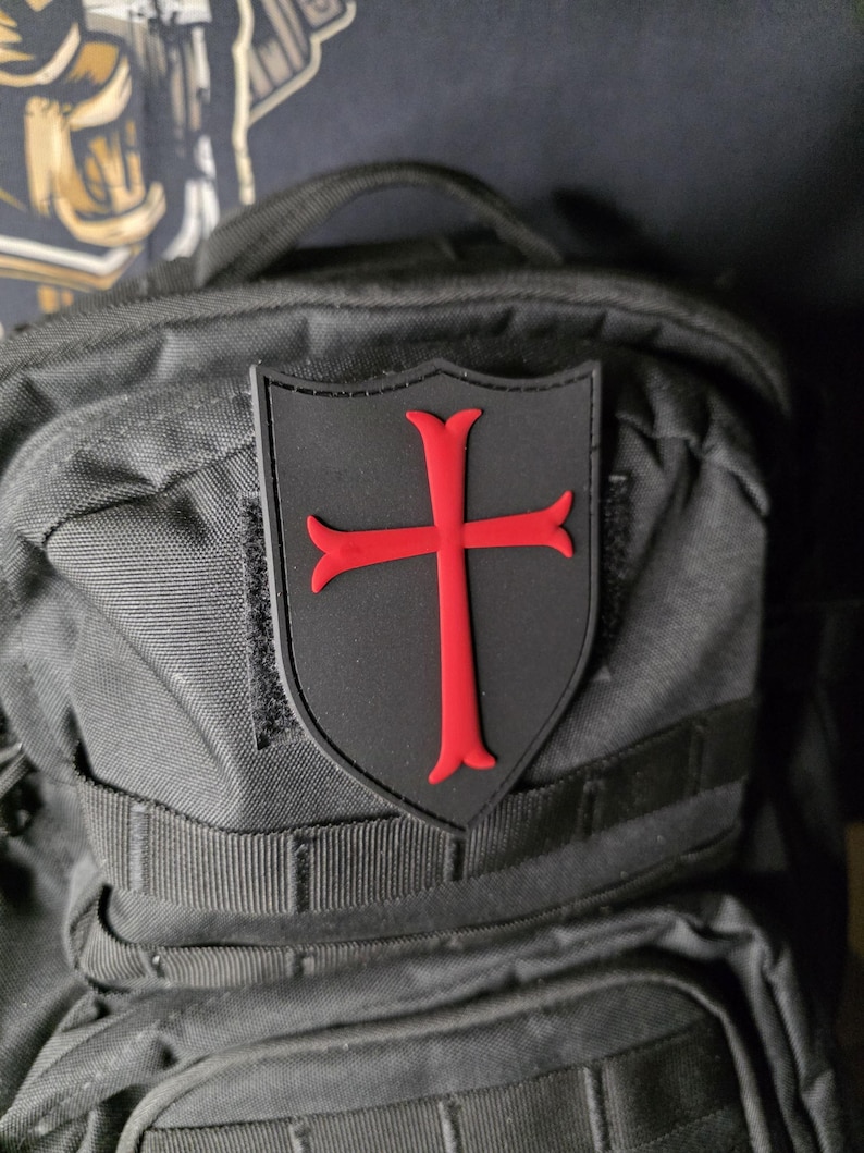 May include: A black patch with a red cross emblem, affixed to a black backpack. The cross is a bold, stylized design, set against a shield-shaped background. The patch is likely made of durable material.