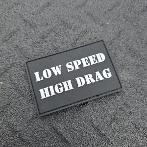 May include: A black rectangular patch with white text that reads "LOW SPEED HIGH DRAG." The patch is likely made of a durable material and is designed to be attached to clothing or gear. The background is a textured, dark gray surface.
