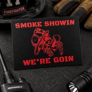 May include: A rectangular black sticker featuring red text that reads SMOKE SHOWIN WE'RE GOIN. The graphic depicts a red skeleton wearing a firefighter helmet and holding a pipe. The design is printed on a matte black surface with sharp, bold lettering.