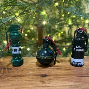 May include: Three novelty grenade ornaments in green and black, with red pull cords. One is cylindrical with white accents, another is spherical, and the third is labeled "M18 SMOKE" with the number "008-831".