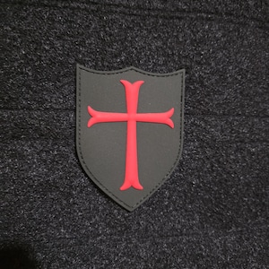 May include: Two patches are shown. The first is a black shield with a red cross. The second is a black rectangle with the words "WE'RE GOIN" in red. Both patches are on a dark gray textured background.